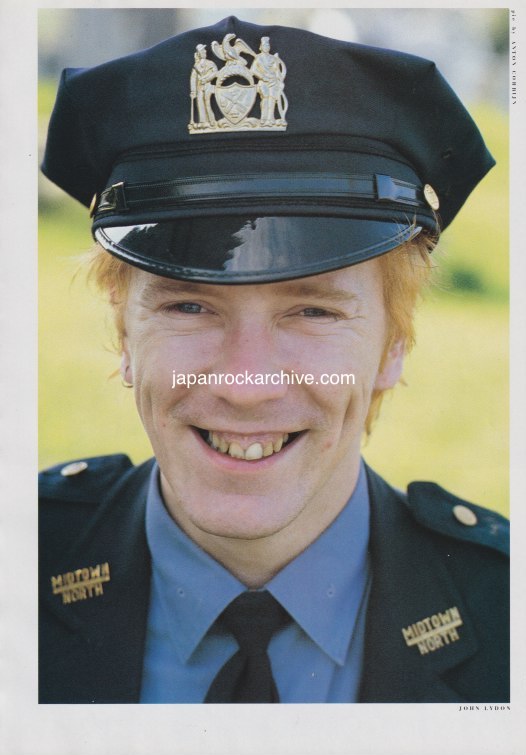 John Lydon of PIL wearing a NYPD Midtown North police uniform with a cap featuring a badge, smiling outdoors.