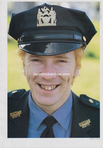 John Lydon of PIL wearing a NYPD Midtown North police uniform with a cap featuring a badge, smiling outdoors.