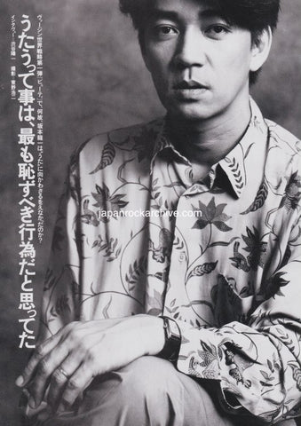 ryuichi sakamoto of ymo wearing a flower patterned shirt sitting.